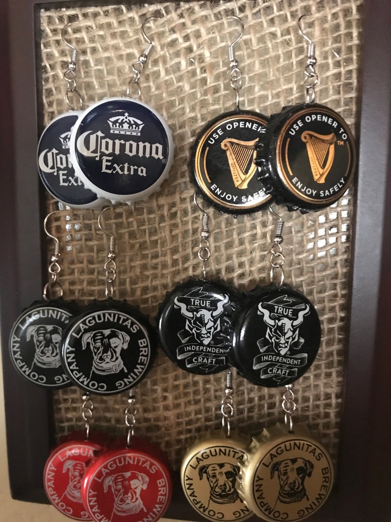 May include: Eight pairs of earrings made from bottle caps. The bottle caps are decorated with various logos and designs, including Corona Extra, Guinness, and Lagunitas Brewing Company. The earrings are silver and have a simple design.