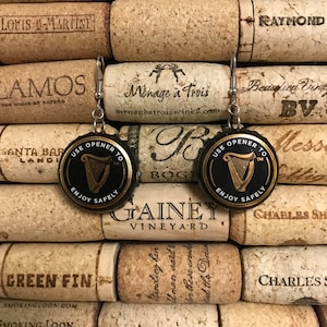 May include: A pair of earrings made from repurposed bottle caps. Each earring features a black and gold bottle cap with a harp design and the text "Use Opener To" and "Enjoy Safely".