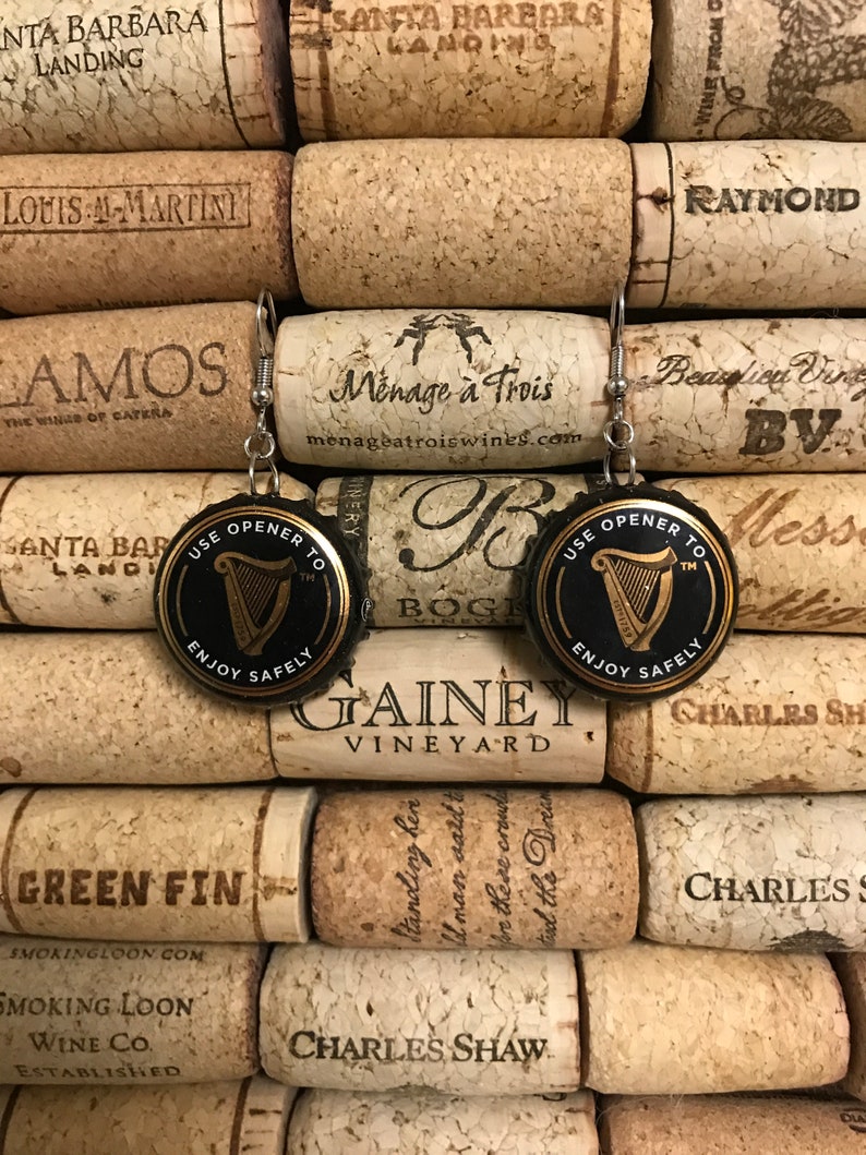 May include: A pair of earrings made from repurposed bottle caps. Each earring features a black and gold bottle cap with a harp design and the text "Use Opener To" and "Enjoy Safely".