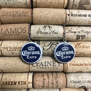 May include: A pair of blue and white Corona Extra bottle cap earrings. The earrings are hanging from cork wine bottle stoppers.