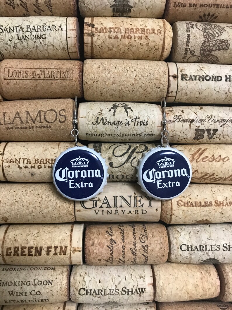May include: A pair of blue and white Corona Extra bottle cap earrings. The earrings are hanging from cork wine bottle stoppers.