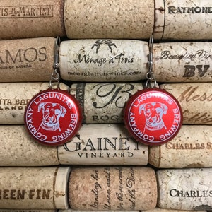 May include: A pair of earrings made from red bottle caps with a white dog silhouette and the text "Lagunitas Brewing Company" on each cap. The bottle caps are attached to silver earring hooks.