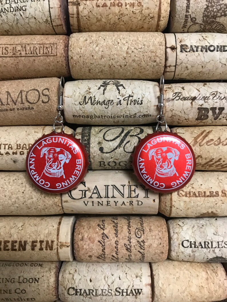 May include: A pair of earrings made from red bottle caps with a white dog silhouette and the text "Lagunitas Brewing Company" on each cap. The bottle caps are attached to silver earring hooks.
