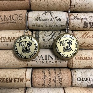 May include: A pair of gold-colored bottle cap earrings with a black and white image of a dog on each cap. The text "Lagunitas Brewing Company" is printed on each cap.