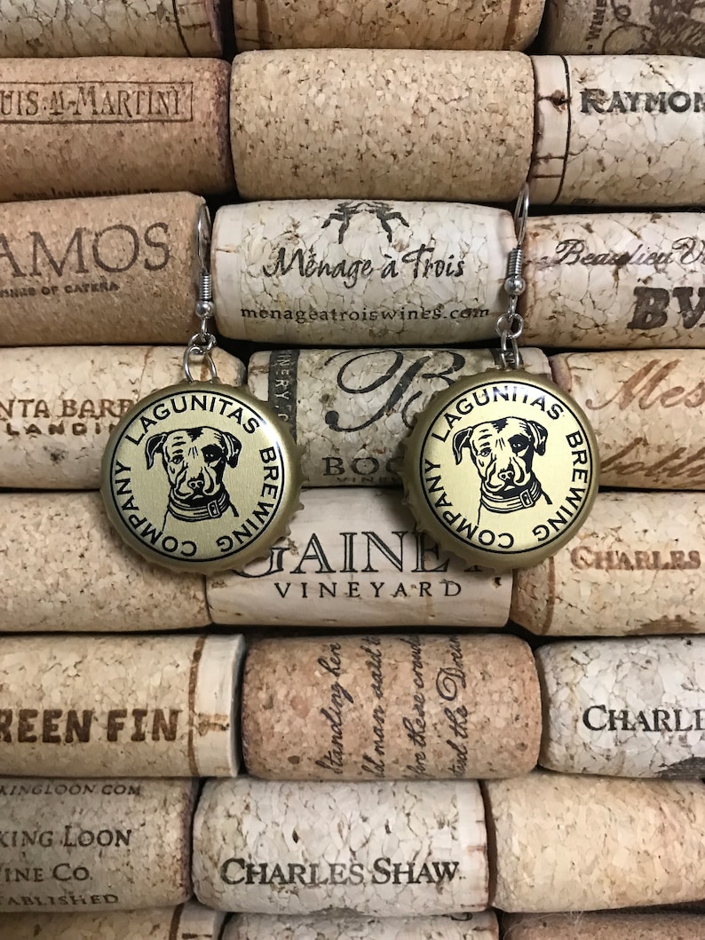 May include: A pair of gold-colored bottle cap earrings with a black and white image of a dog on each cap. The text "Lagunitas Brewing Company" is printed on each cap.