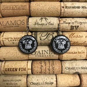 May include: A pair of earrings made from black bottle caps with a white logo that says "Lagunitas Brewing Company" and a cartoon dog wearing a crown. The bottle caps are surrounded by cork wine bottle stoppers.