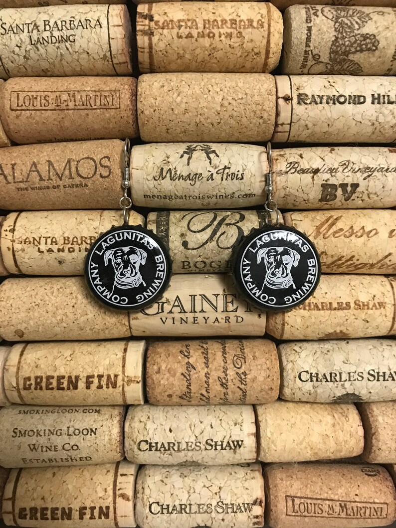 May include: A pair of earrings made from black bottle caps with a white logo that says "Lagunitas Brewing Company" and a cartoon dog wearing a crown. The bottle caps are surrounded by cork wine bottle stoppers.