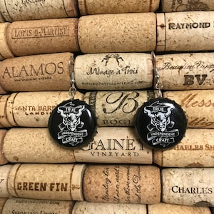 May include: A pair of earrings featuring black and white bottle caps with a graphic of a demon and the text "TRUE INDEPENDENT CRAFT". The bottle caps are attached to silver earring hooks.