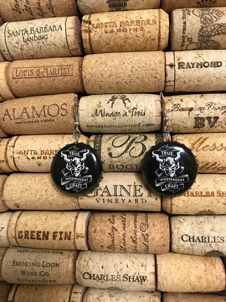 May include: A pair of earrings featuring black and white bottle caps with a graphic of a demon and the text "TRUE INDEPENDENT CRAFT". The bottle caps are attached to silver earring hooks.