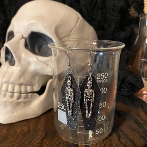 May include: A pair of black glitter coffin-shaped earrings with silver skeleton charms hanging from silver hooks.
