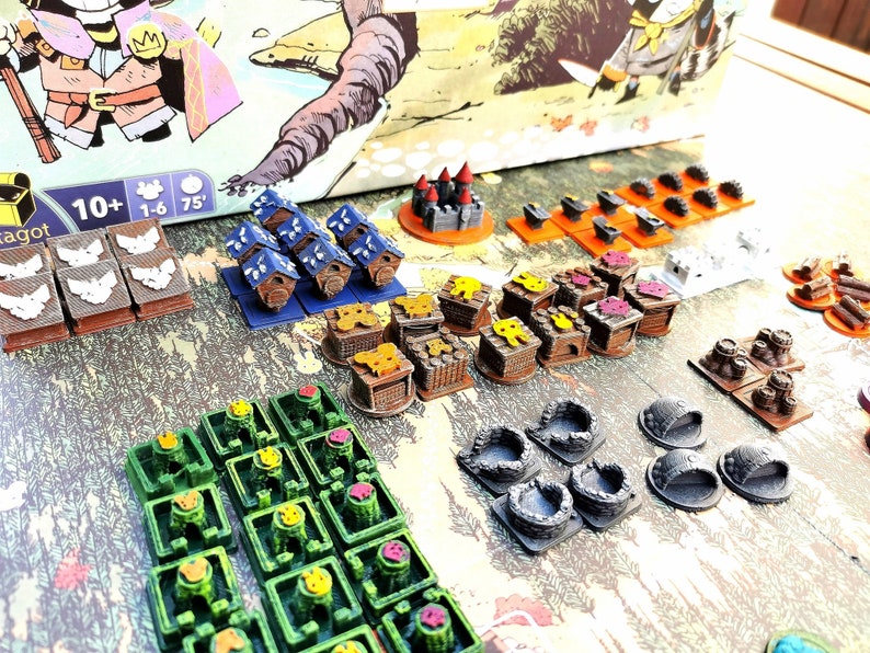 May include: A collection of colorful wooden game pieces for a board game. The pieces include houses, castles, and other structures. The pieces are made of wood and painted in various colors, including blue, green, yellow, red, and brown. The pieces are arranged on a table, and there are several other game pieces in the background.