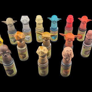 Könnte beinhalten: Eine Sammlung von 18 kleinen Farbflaschen mit verschiedenen Figuren auf den Kappen. Die Flaschen sind in einem Halbkreis auf einem schwarzen Hintergrund angeordnet. Zu den Figuren gehören Yoda, ein Mandalorianer und ein Stormtrooper.