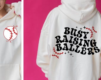 Puede incluir: Sudadera blanca de gran tamaño con un gráfico de béisbol en la parte delantera y el texto "Busy Raising Ballers" en la parte posterior.