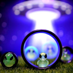 Può includere: Un'immagine stravagante con gioielli a tema alieno. Il punto focale è una figura aliena verde trasparente all'interno di un cerchio con bordo nero. Altri pezzi includono una faccia aliena verde e altri oggetti a tema alieno, il tutto sullo sfondo di un UFO e un cielo blu stellato.