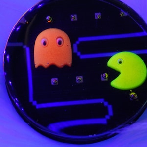 May include: A black and blue circular coaster with a Pac-Man design. The coaster features a yellow Pac-Man and an orange ghost.