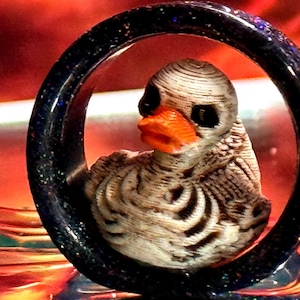 May include: A unique, dark blue ear gauge with a skeletal duck design. The duck has an orange beak and detailed rib cage. The gauge is set against a vibrant red and orange background, creating a striking contrast.