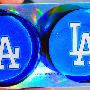 May include: Two blue round glass coasters with a white LA logo on each. The coasters are on a holographic background.