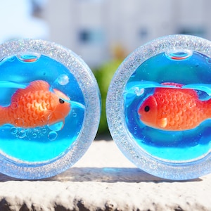 May include: Two clear glass ornaments with a silver glitter rim, each containing a small orange fish swimming in blue resin.