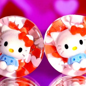 Può includere: Un paio di dilatatori per orecchie in acrilico trasparente con un design Hello Kitty. I dilatatori sono riempiti con cuori rossi e bianchi e glitter.