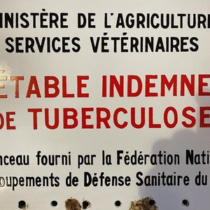 Op de afbeelding: Wit emaille bord met zwarte en rode tekst. Het bord luidt "Ministère de l'Agriculture Services Vétérinaires Étable Indemne de Tuberculose". Het bord bevat ook de tekst "Panonceau fourni par la Fédération Nationale des Groupements de Défense Sanitaire du Bétail".