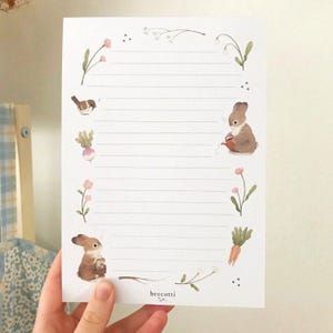 May include: A white lined paper with a floral border and illustrations of bunnies and birds. The paper has the text "beccotti" at the bottom.