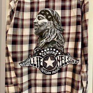 May include: A black and white flannel shirt with a painted portrait of Willie Nelson and the text "Genuine Outlaw Music Willie Nelson".