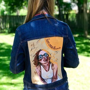 May include: A denim jacket with a painted design on the back. The design features a woman wearing sunglasses and a white shirt with the text "Here comes the sun" in yellow.