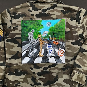 May include: A camouflage jacket with a painted image of Star Wars characters walking across a crosswalk. The characters include R2-D2, C-3PO, BB-8, and a Stormtrooper.