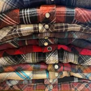 Pode incluir: Uma pilha de camisas de flanela dobradas em várias cores e padrões xadrez. As camisas apresentam fechos de botões e uma variedade de tons, incluindo vermelho, preto, castanho e azul. A imagem mostra a textura e os detalhes do tecido.