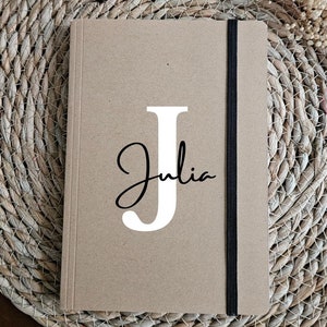 Puede incluir: Un cuaderno marrón personalizado con el nombre "Julia" y una "J" blanca en la portada. Tiene una banda elástica negra. Una vela en forma de flor y flores secas en el fondo.