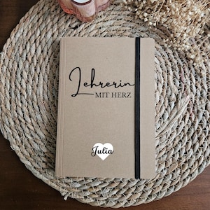Könnte beinhalten: Ein braunes Notizbuch mit dem Text "Lehrerin mit Herz" und einem weißen Herz mit dem Namen "Julia" darauf gedruckt. Das Notizbuch liegt auf einer geflochtenen Stroh-Unterlage mit einer weißen Blumenkerze.