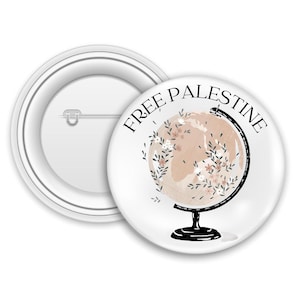 Op de afbeelding: Witte button met de tekst "FREE PALESTINE" in zwart, rondom een wereldbolontwerp. De wereldbol is licht perzikkleurig met bloemaccenten en een zwarte standaard. Een tweede button is zichtbaar op de achtergrond.