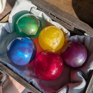 May include: Five colorful, spherical orbs in a wooden box. The orbs are green, yellow, blue, red, and purple, each with a shimmering, iridescent effect. The box is lined with a light blue fabric.