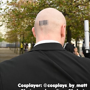 May include: A bald head featuring a black barcode tattoo with the numbers 640509 040147 printed below it. The individual is wearing a black suit jacket with a white shirt collar visible underneath and is holding a silver handgun in a gloved hand.