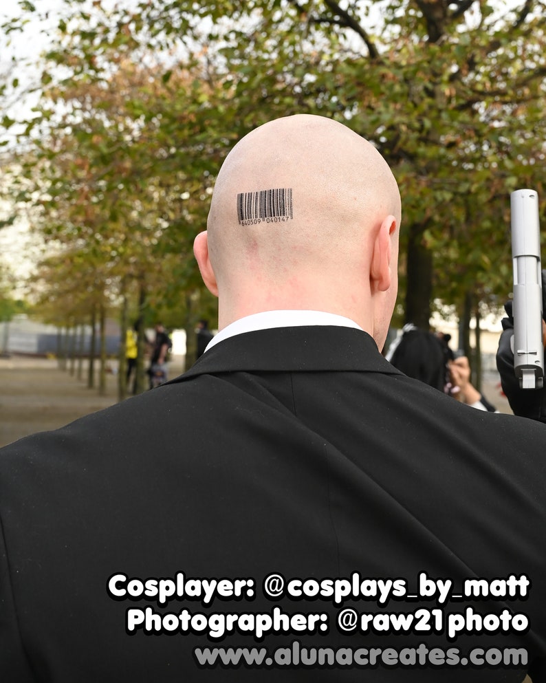May include: A bald head featuring a black barcode tattoo with the numbers 640509 040147 printed below it. The individual is wearing a black suit jacket with a white shirt collar visible underneath and is holding a silver handgun in a gloved hand.