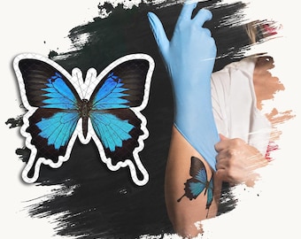 May include: A temporary tattoo design featuring a vibrant blue and black butterfly with elongated lower wings. The butterfly is displayed as a standalone graphic and also shown applied to skin, with a light blue glove visible nearby.