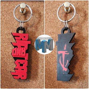 May include: Two keychains with silver key rings. One keychain is black with red text that reads "RIP AND TEAR". The other keychain is black with a red symbol. Both keychains are attached to a corkboard.