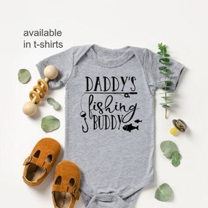 May include: Grey baby growsuit with the text "Daddy's fishing buddy" in black lettering. A fishing hook and two fish are also included in the design. The growsuit is surrounded by a wooden rattle, brown baby shoes, and green eucalyptus leaves.