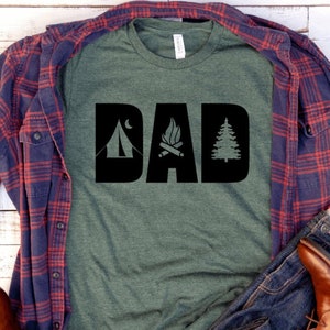 Pode incluir: Uma camiseta verde com um gr&aacute;fico preto que diz "DAD" com &iacute;cones de barraca, fogueira e pinheiro. A camiseta est&aacute; sobreposta a uma camisa xadrez azul e vermelha e uma cal&ccedil;a jeans azul.