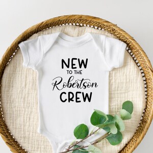 Peut inclure: Un body bébé blanc avec un texte noir qui dit "New to the Robertson Crew". Le body est posé sur un panier en osier avec une brindille de verdure.