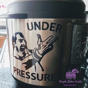 May include: A silver Instant Pot with a black vinyl decal that says "Under Pressure" with a silhouette of a person singing into a microphone. The decal also includes a hand making the "rock on" hand gesture. The Instant Pot is sitting on a black countertop. The decal is from Purple Zebra Crafts, Custom Vinyl Products.