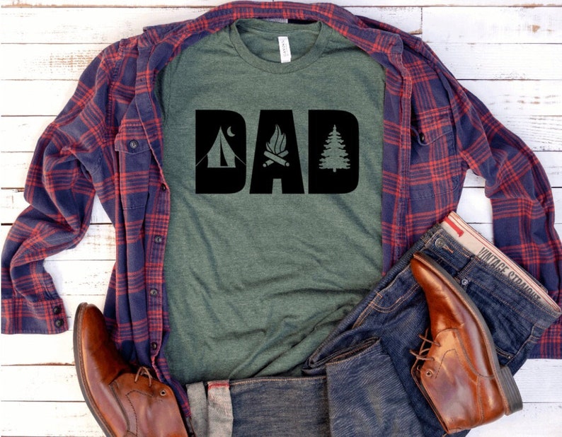 Pode incluir: Uma camiseta verde com um gr&aacute;fico preto que diz "DAD" com &iacute;cones de barraca, fogueira e pinheiro. A camiseta est&aacute; sobreposta a uma camisa xadrez azul e vermelha e uma cal&ccedil;a jeans azul.