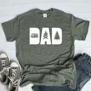 Pode incluir: Uma camiseta verde com a palavra "DAD" em letras brancas. As letras s&atilde;o decoradas com um trailer, uma fogueira e um pinheiro.