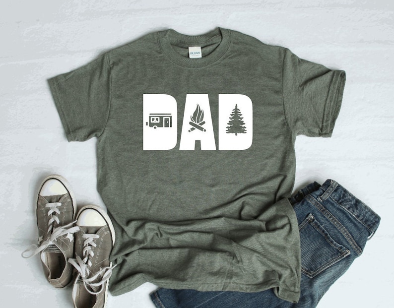 Pode incluir: Uma camiseta verde com a palavra "DAD" em letras brancas. As letras s&atilde;o decoradas com um trailer, uma fogueira e um pinheiro.