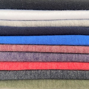 Pode incluir: Uma pilha de t-shirts dobradas em v&aacute;rias cores, incluindo preto, branco, cinza, azul, vermelho e verde.