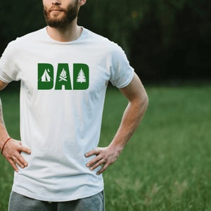 Pode incluir: Camiseta branca com a palavra "DAD" em letras verdes. Cada letra &eacute; decorada com um &iacute;cone tem&aacute;tico de acampamento: uma barraca, uma fogueira e um pinheiro.
