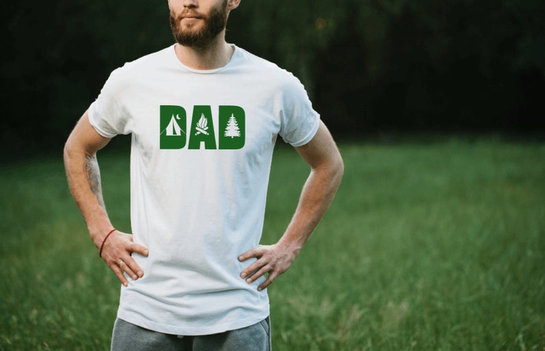 Pode incluir: Camiseta branca com a palavra "DAD" em letras verdes. Cada letra &eacute; decorada com um &iacute;cone tem&aacute;tico de acampamento: uma barraca, uma fogueira e um pinheiro.