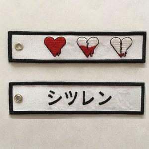 May include: Two white fabric keychains with black trim. The top keychain has three embroidered red hearts, two of which are broken in half. The bottom keychain has the Japanese word "シツレン" embroidered in black.