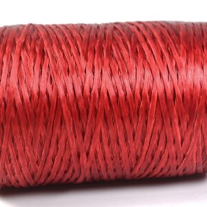 May include: A spool of red raffia ribbon. The ribbon is tightly wound around a cardboard core, creating a cylindrical shape. The red color is vibrant and consistent throughout the spool, suitable for crafting and gift wrapping.