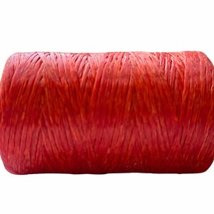 May include: A cylindrical spool of vibrant red raffia ribbon, tightly wound with a visible fibrous texture. The material displays a consistent, saturated red hue throughout the entire roll, showcasing a natural, slightly matte finish with distinct vertical strands.